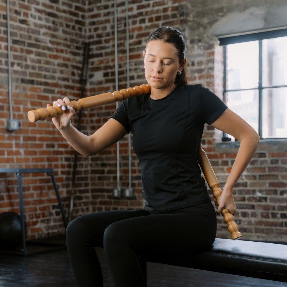 Woman holding a wooden Body Lever massage tool in a gym setting with brick walls.