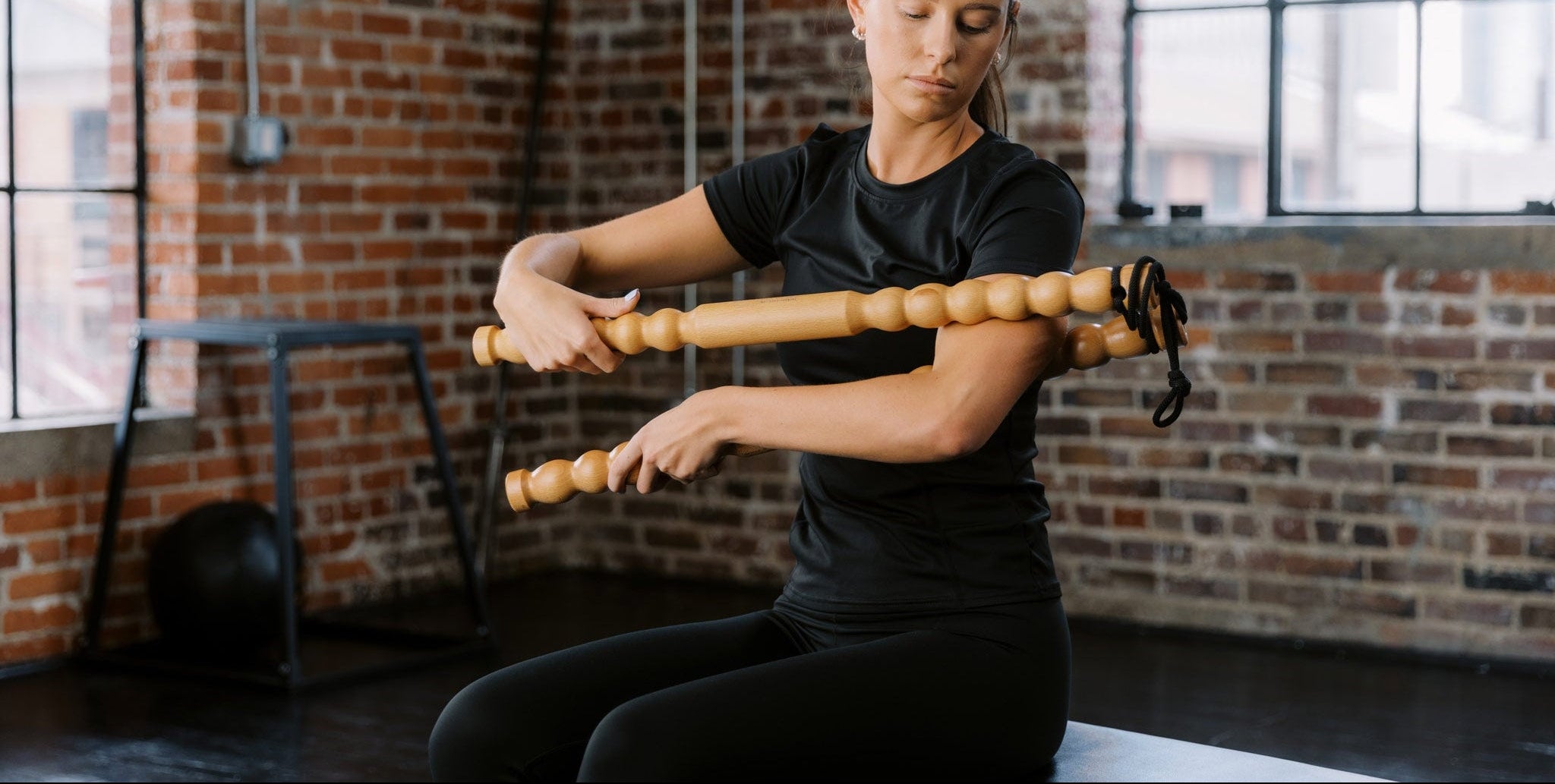 Woman using a wooden muscle massage tool in a gym setting with brick walls and large windows.