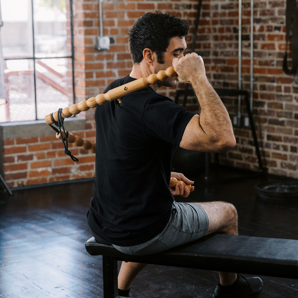 Man using a wooden fascia massage sticks on him back in a gym setting with brick walls.