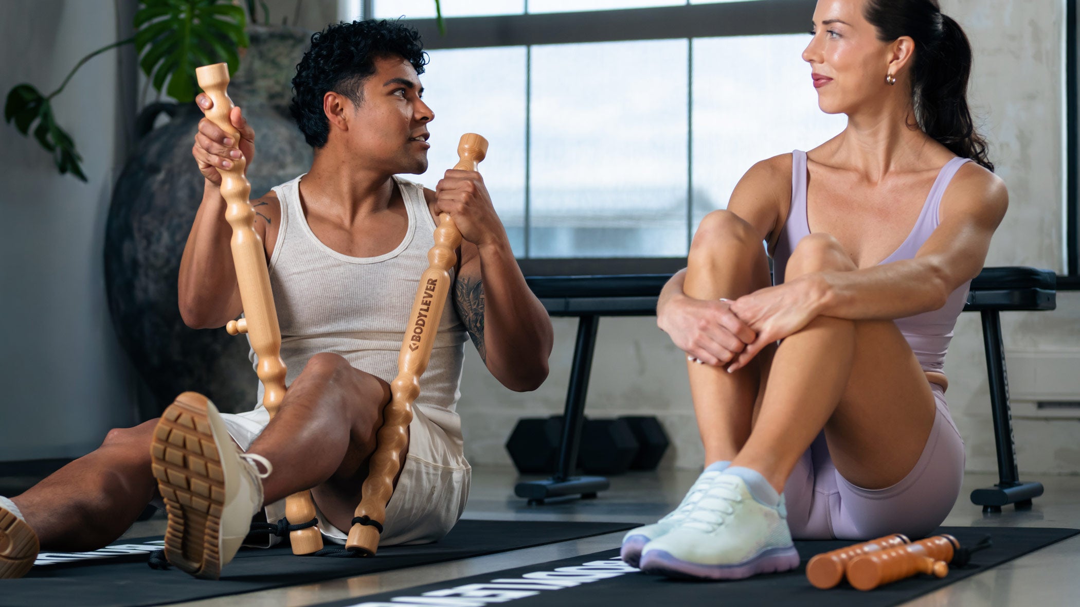 Two people in workout attire sitting on exercise mats in a bright gym, each holding wooden massage handles. One person holds the handles upright while the other sits with legs bent, showing the product in use in a real environment.