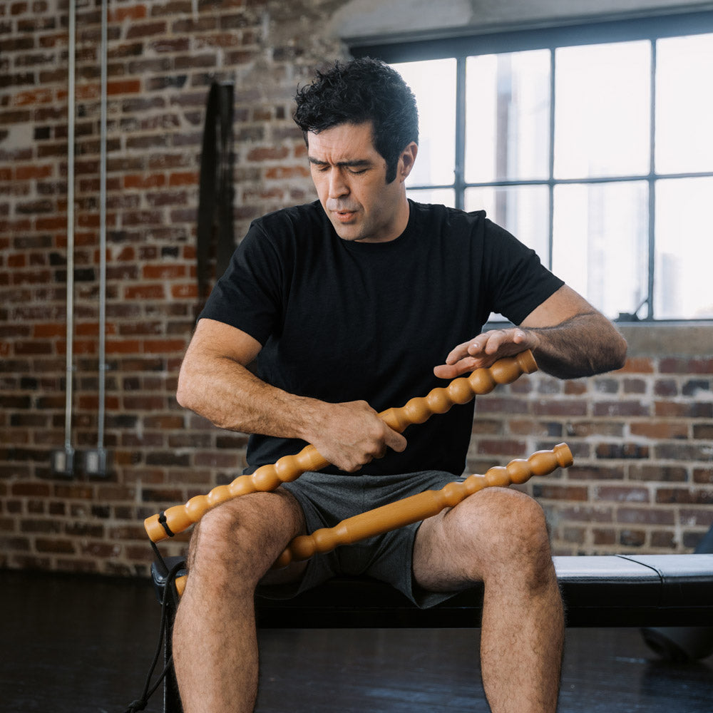 Man holding wooden massage sticks in a room with brick walls and large windows.