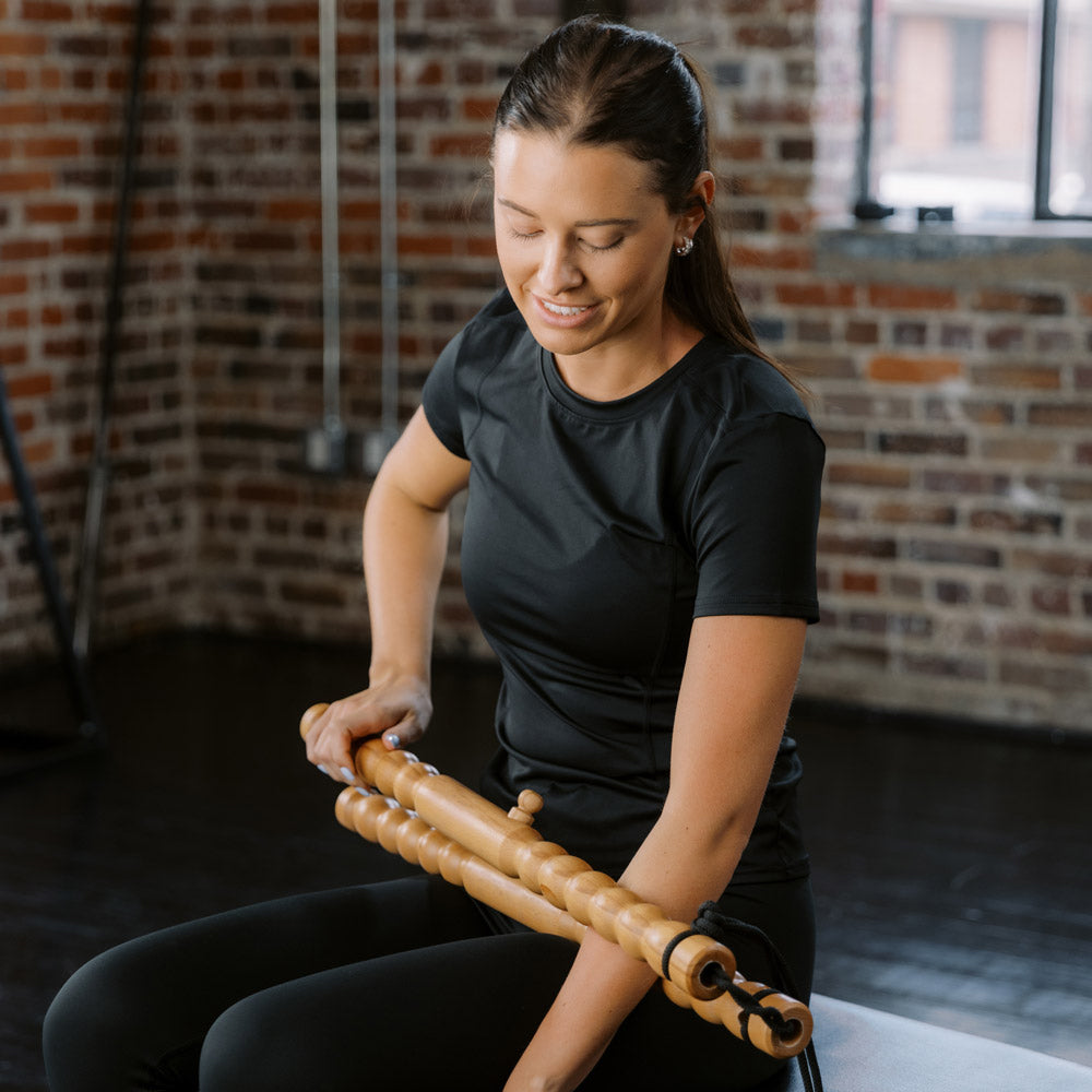 Woman using a wooden massage tool roller tool in a gym setting with brick walls.